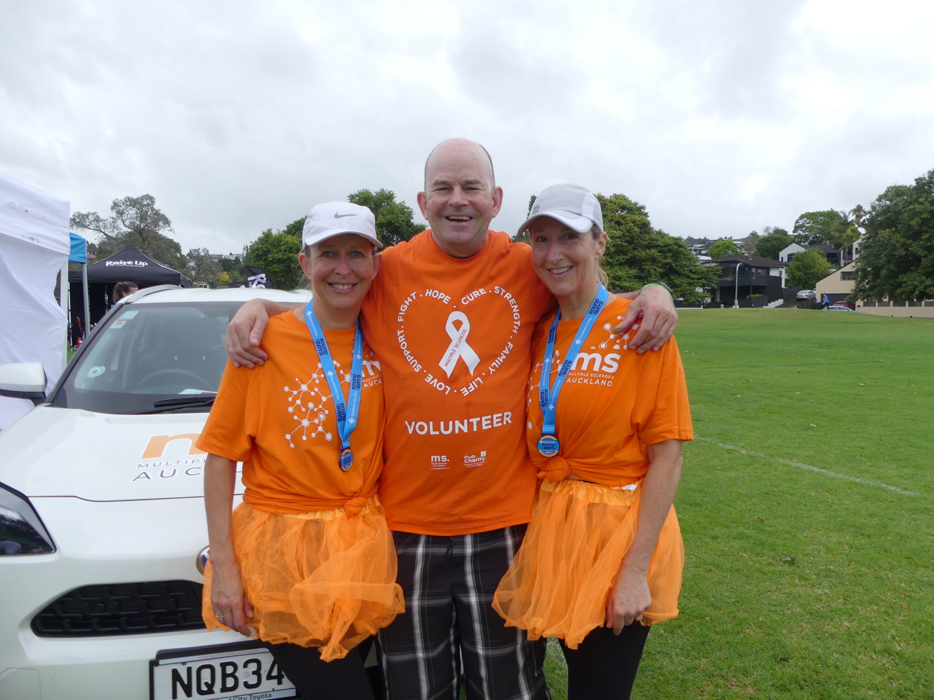 Andrew Coe with a volunteering orange t-shirt on with his sisters in orange t-shirts, outside after Round the Bays 2024